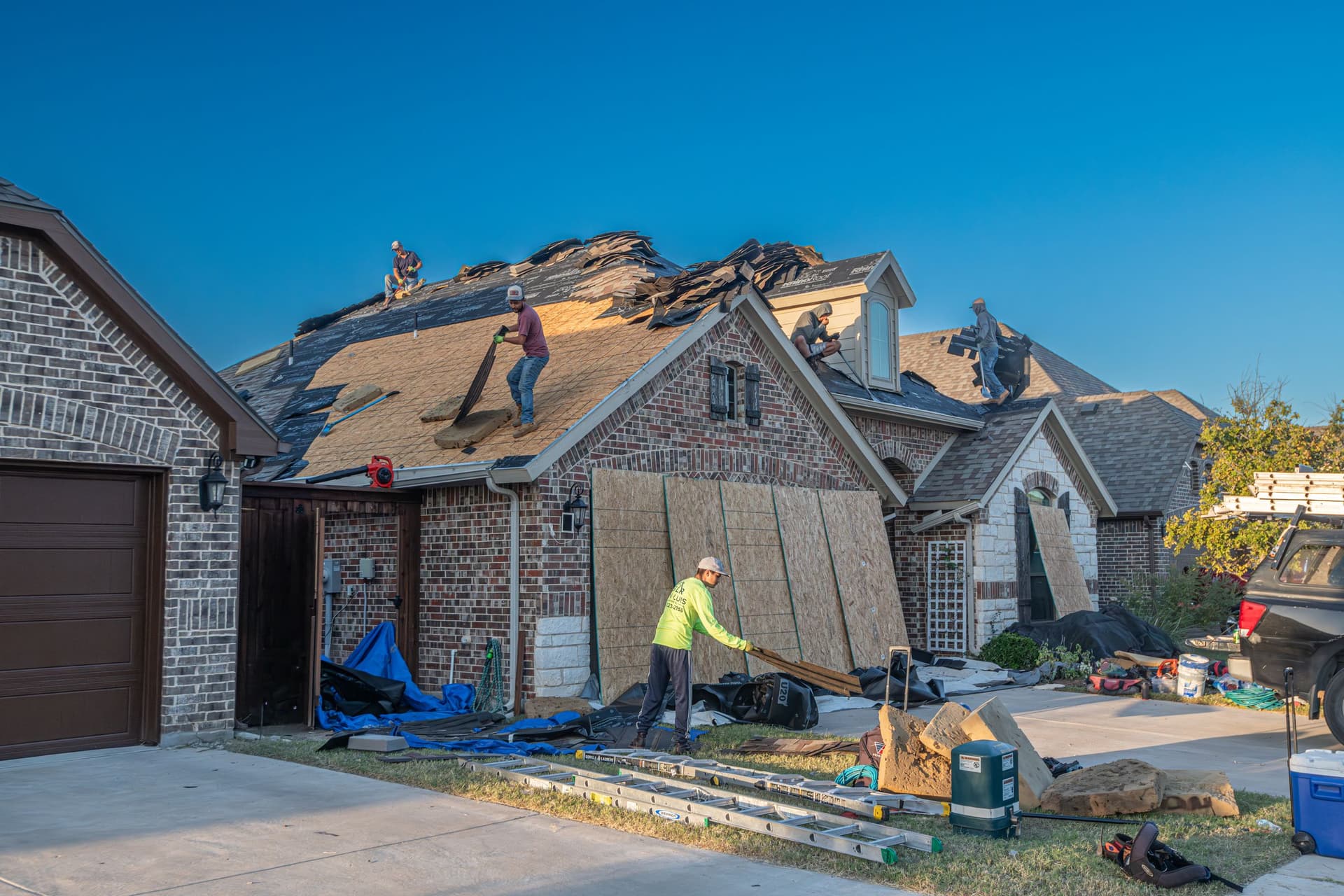 Roofing crew tearing off shingles on a brick home, with new decking sheets leaning against the wall