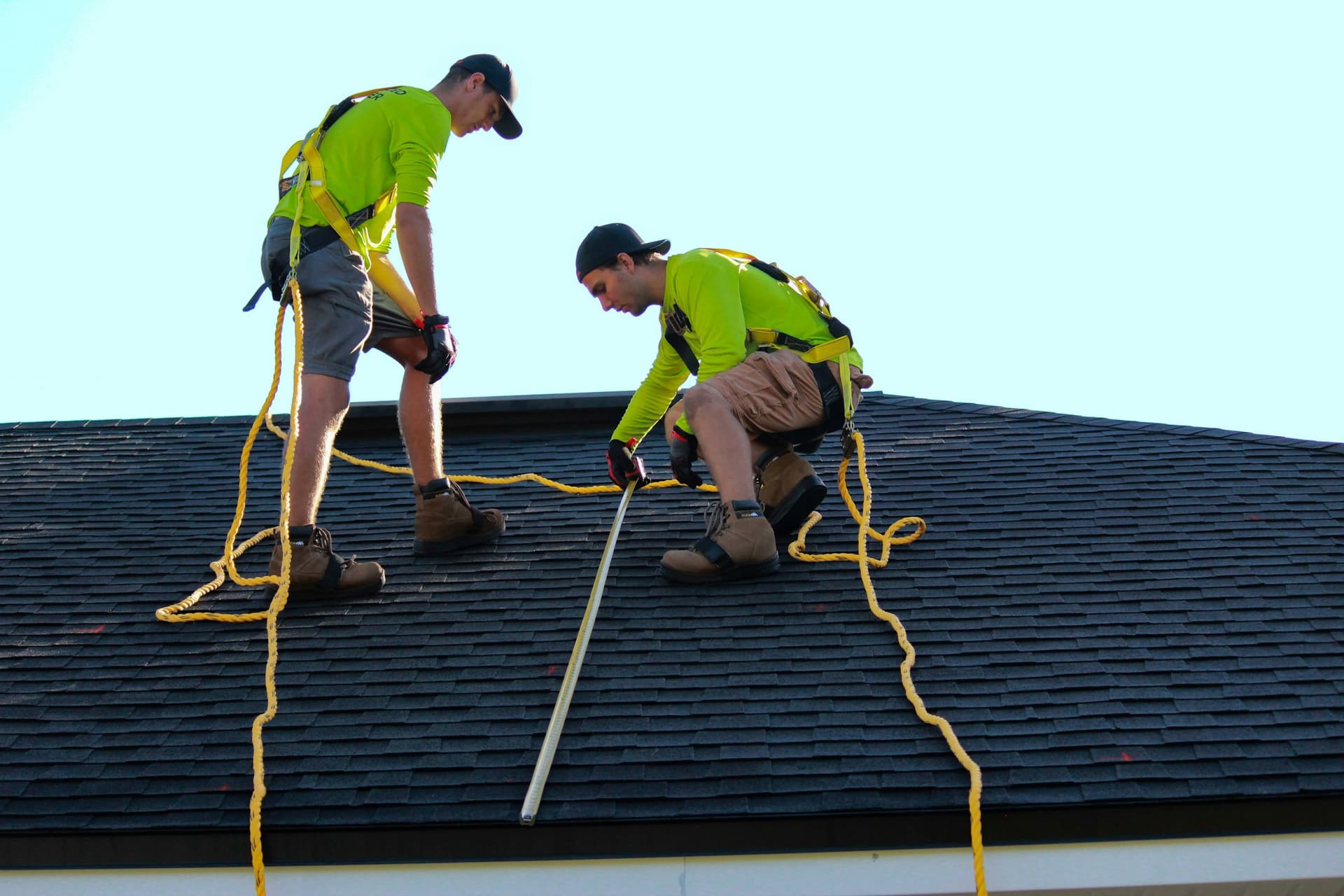 Two roofers in fall protection running a tape measure down an asphalt shingle roof